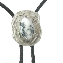Load image into Gallery viewer, A close-up of a bolo tie necklace featuring a textured, organic-shaped silver pendant with a dendritic opal gem in the center, suspended from black braided cords against a white background.