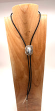 Load image into Gallery viewer, A black braided bolo-style necklace with a polished sterling silver and dendritic opal gem displayed on a light wood neck mannequin against a white background.