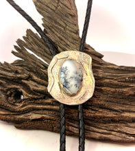 Load image into Gallery viewer, A close-up of a rustic silver-toned bolo tie necklace with a dendritic opal gem, suspended over a piece of weathered driftwood.