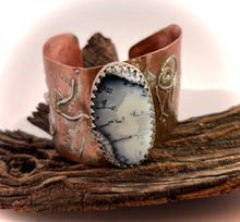 Load image into Gallery viewer, A handmade cuff bracelet made of copper and silver with engraved designs, featuring a large oval dendritic opal gem, displayed on a piece of weathered wood.