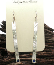 Load image into Gallery viewer, A pair of long, textured sterling silver dangle earrings with faceted 4mm sapphire gems displayed on a cream card over a piece of dark wood.