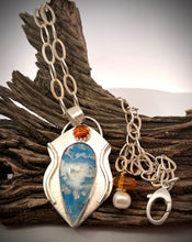 Load image into Gallery viewer, A silver pendant necklace displayed on a piece of weathered driftwood. The teardrop shaped pendant features a blue and white cloud pattern , a small round Baltic amber gem and a dangling charm with a white pearl and amber bead.