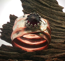 Load image into Gallery viewer, A handmade ring with a textured, hammered copper and silver band featuring a central faceted garnet red gemstone in a crown-like setting, displayed on a piece of weathered wood.